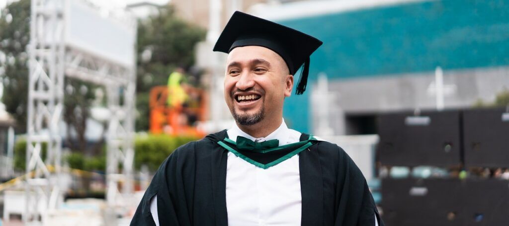 Male student in graduation cap and gown outside smiling