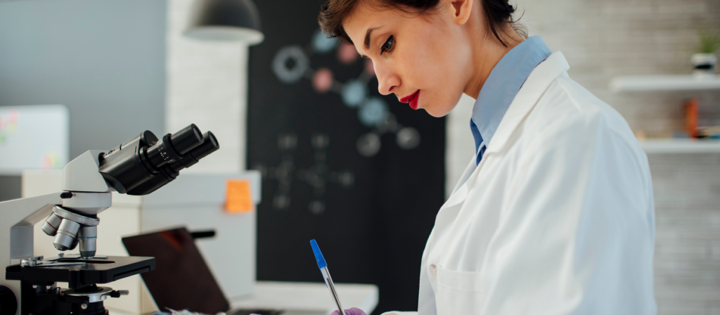 Woman in lab coat near microscope writing down results